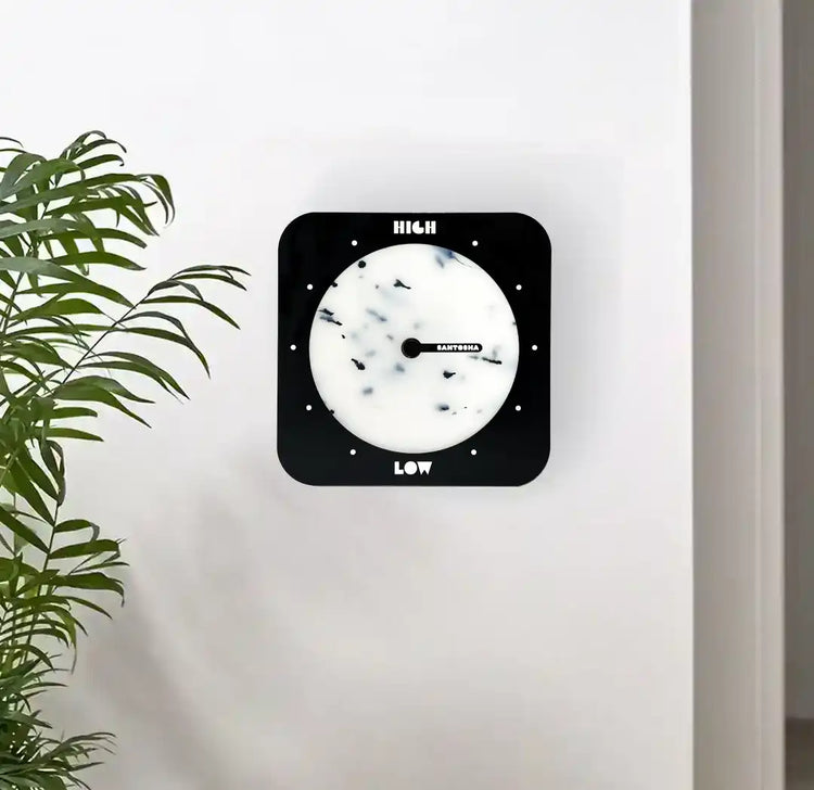 Horloge des marées carrée à bords arrondis noir et blanche accrochée au mur avec une plante verte en avant plan 