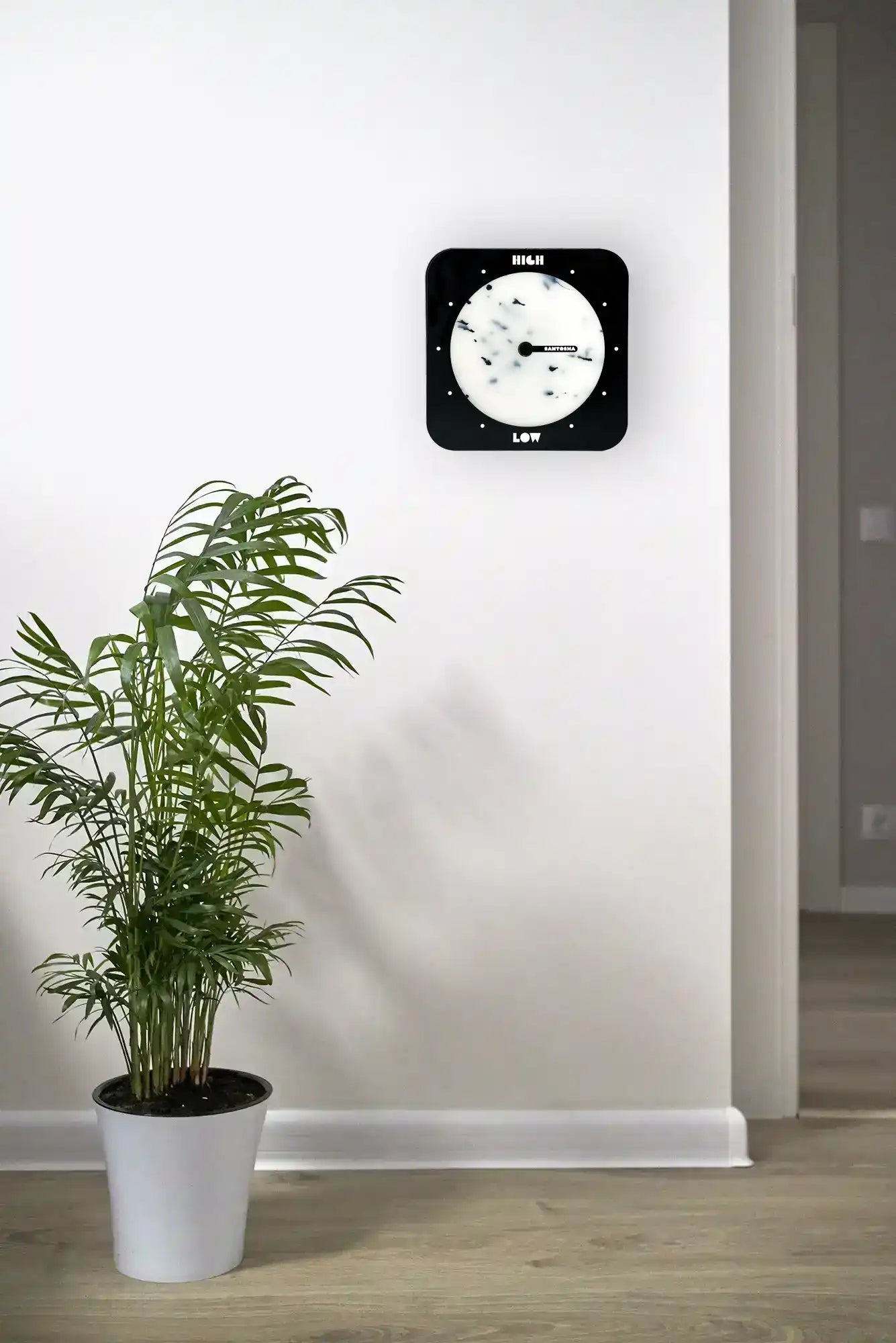 Horloge des marées carrée à bords arrondis noir et blanche accrochée au mur avec une plante verte en avant plan 